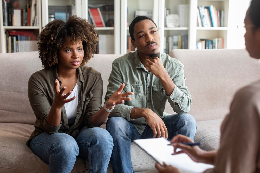 Two adults on a beige couch in a counseling session, one gesturing as the other listens while a therapist takes notes.