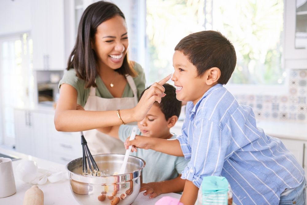 Mom and two kids bake in a bright kitchen; the boy leans toward a mixing bowl as the mom smiles nearby.