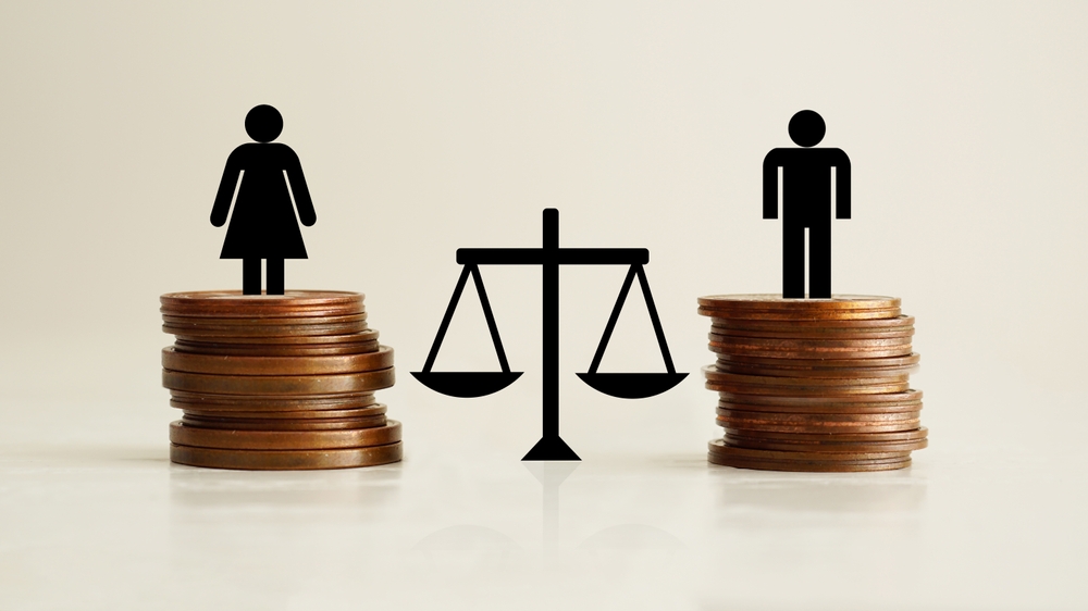 Silhouettes of a woman on a coin stack and a man on another coin stack with a balance scale between them, illustrating the gender pay gap side-by-side the scales suggest imbalance.