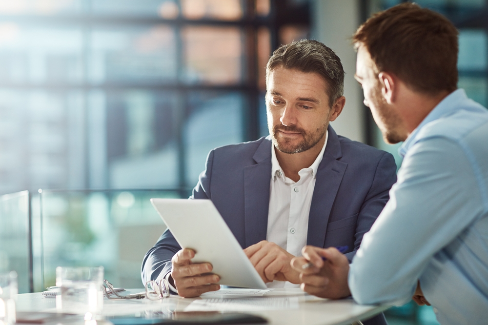 Two businessmen review a tablet and discuss documents at a bright office conference table.