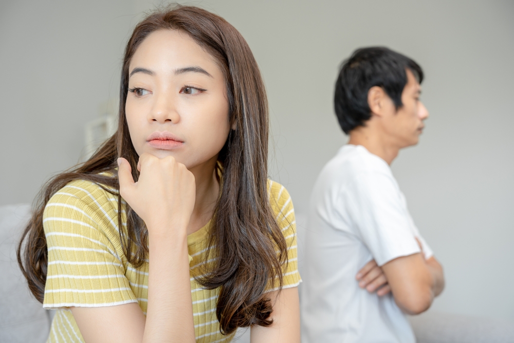 Woman in a yellow striped shirt rests chin on hand, looking away; man in a white shirt stands with arms crossed in the background, suggesting tension.