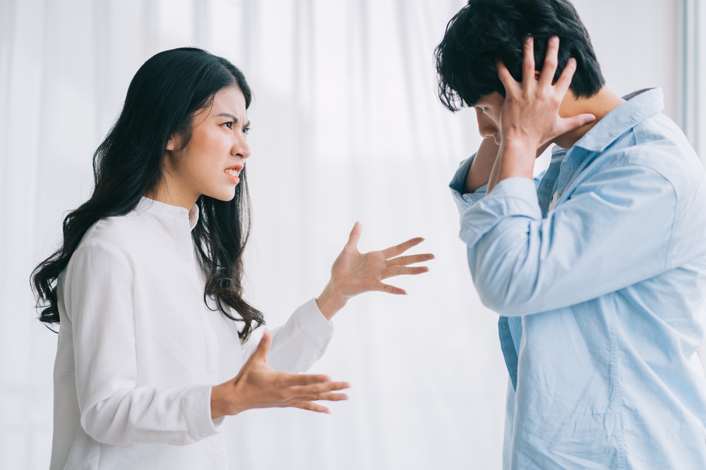 Woman in white gestures angrily at a man who covers his ears during a heated disagreement indoors.