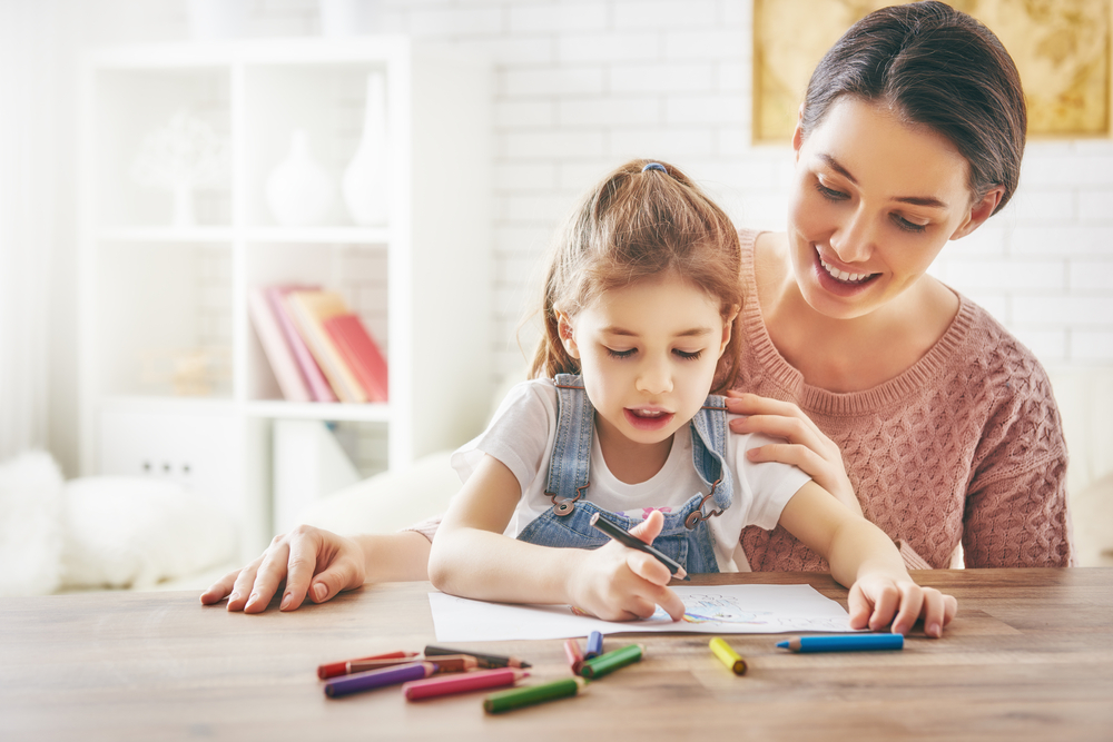 Mother guides her daughter as she draws with crayons at a wooden table in a bright room.